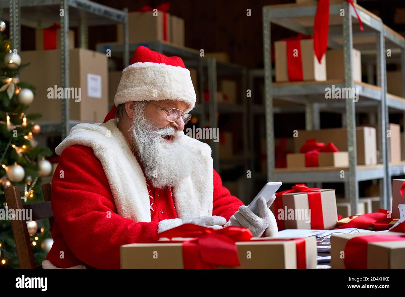 Happy Santa Claus holding cell phone using mobile app sitting at workshop table. Stock Photo