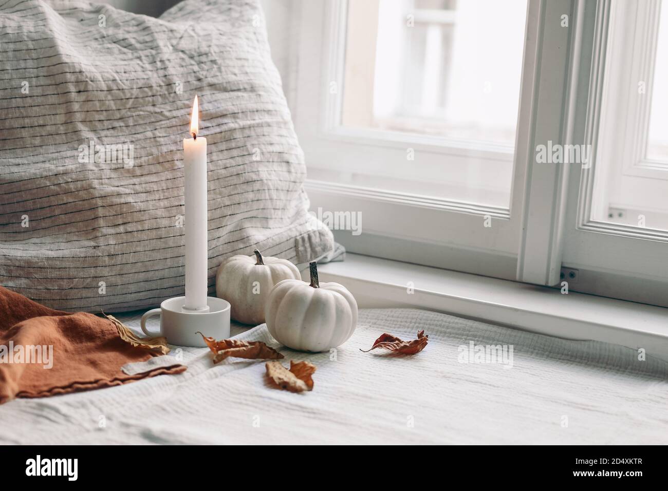 Autumn still life. Burning candle in white ceramic candleholder. Dry beech leaves and linen pillow near window. Moody composition with white pumpkins Stock Photo
