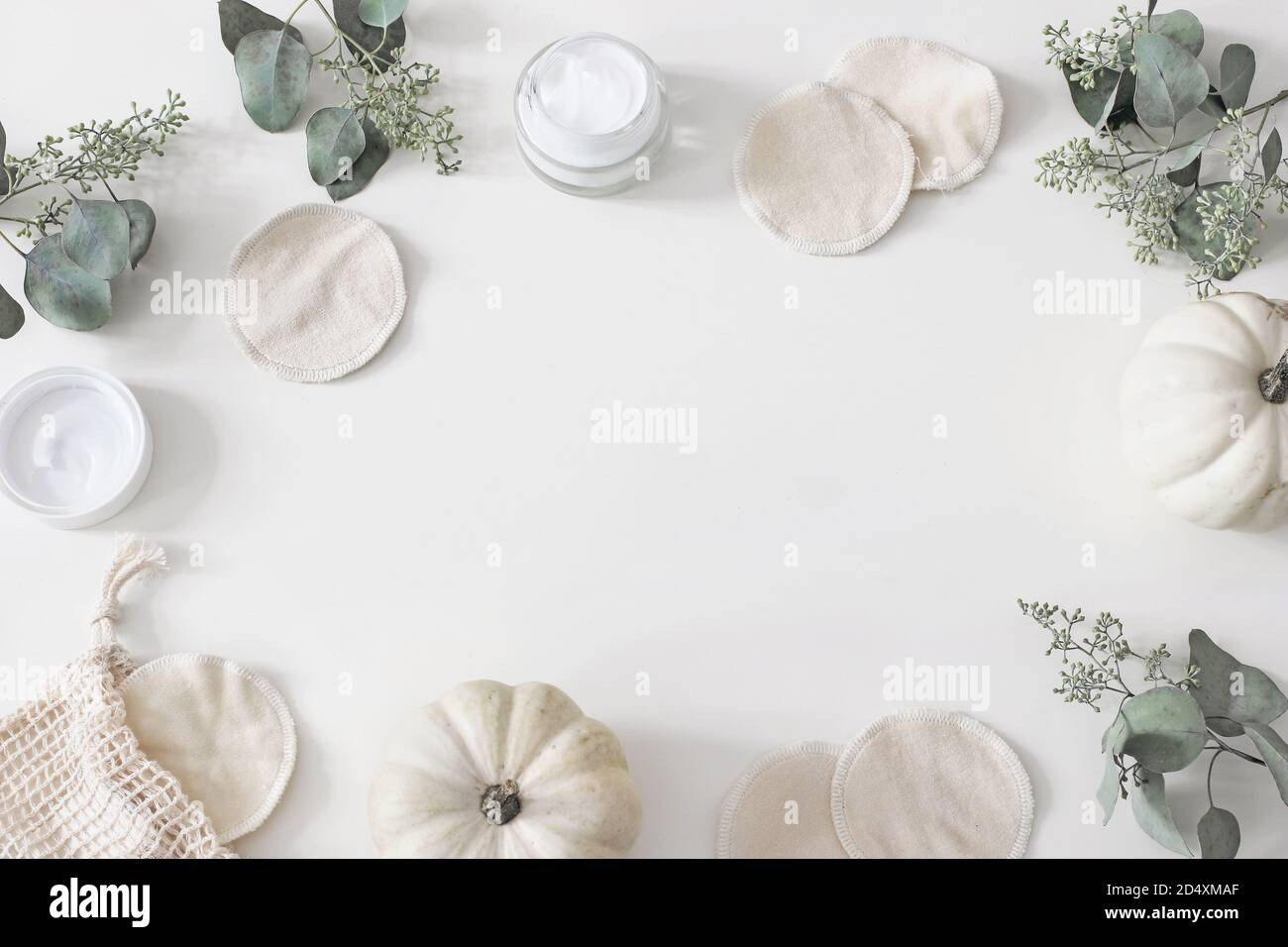 Decorative cosmetics frame. Organic cotton reusable round pads for make up removal. Eucalyptus branches, pumpkins.and moisturizer on white table Stock Photo