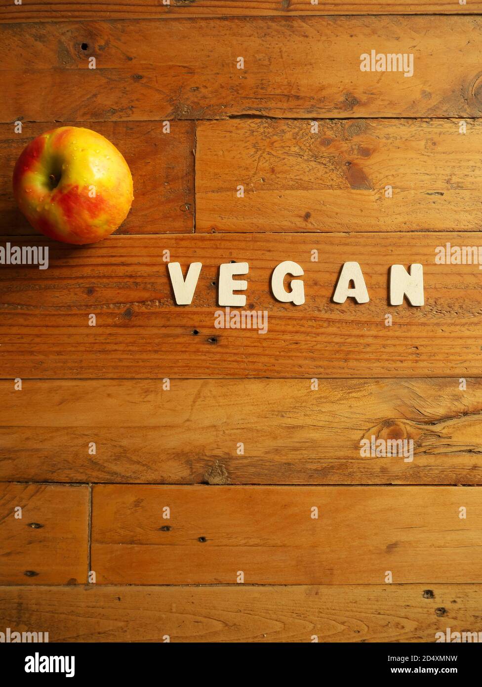 Word 'VEGAN' in white letters on a wooden background with an apple Stock Photo