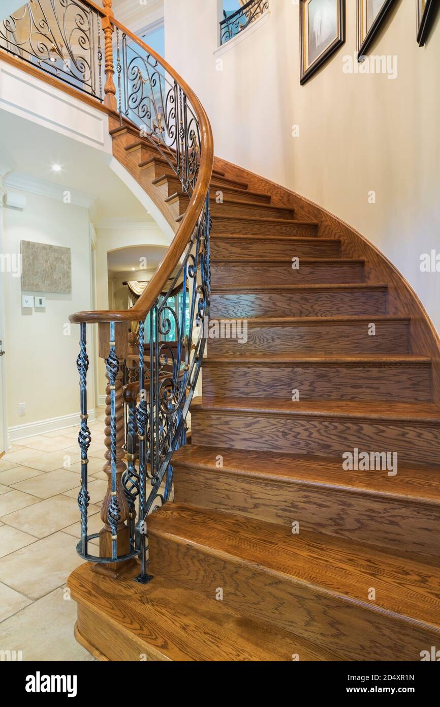 Spiraling wooden staircase with wrought iron baluster leading to upstairs floor inside a luxurious residential home. Stock Photo