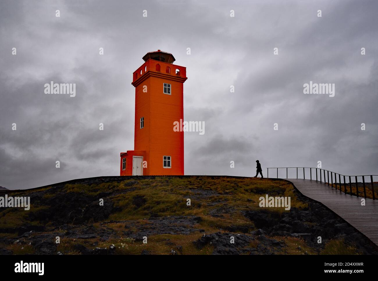 A person walks along a path through lava fields leading up to the orange Svortuloft Lighthouse on the Snaefellsnes Peninsular in Western Iceland. Stock Photo