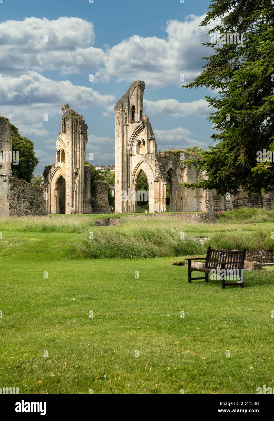 Postcard view of Glastonbury Abbey ruins with green parkland and resting bench in foreground. Blue sky day with white clouds. No people. Copy space. Stock Photo