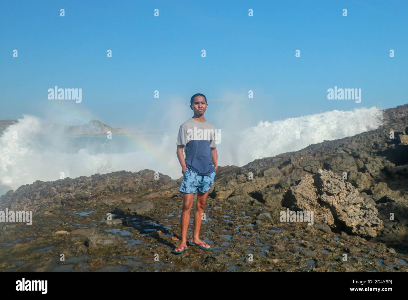 Waves hitting round rocks and splashing. A young man stands on a rocky shore and the waves crash against a cliff Stock Photo