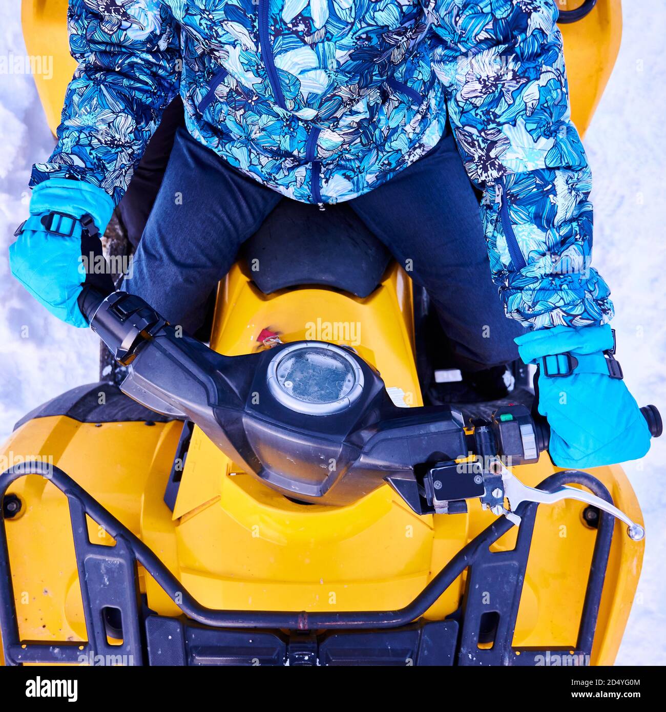 Cropped close-up snapshot of person in blue winter suit and mittens,  sitting on yellow ATV bike holding helm in winter, top view. Concept of  winter entertainment, activities and quad biking Stock Photo -