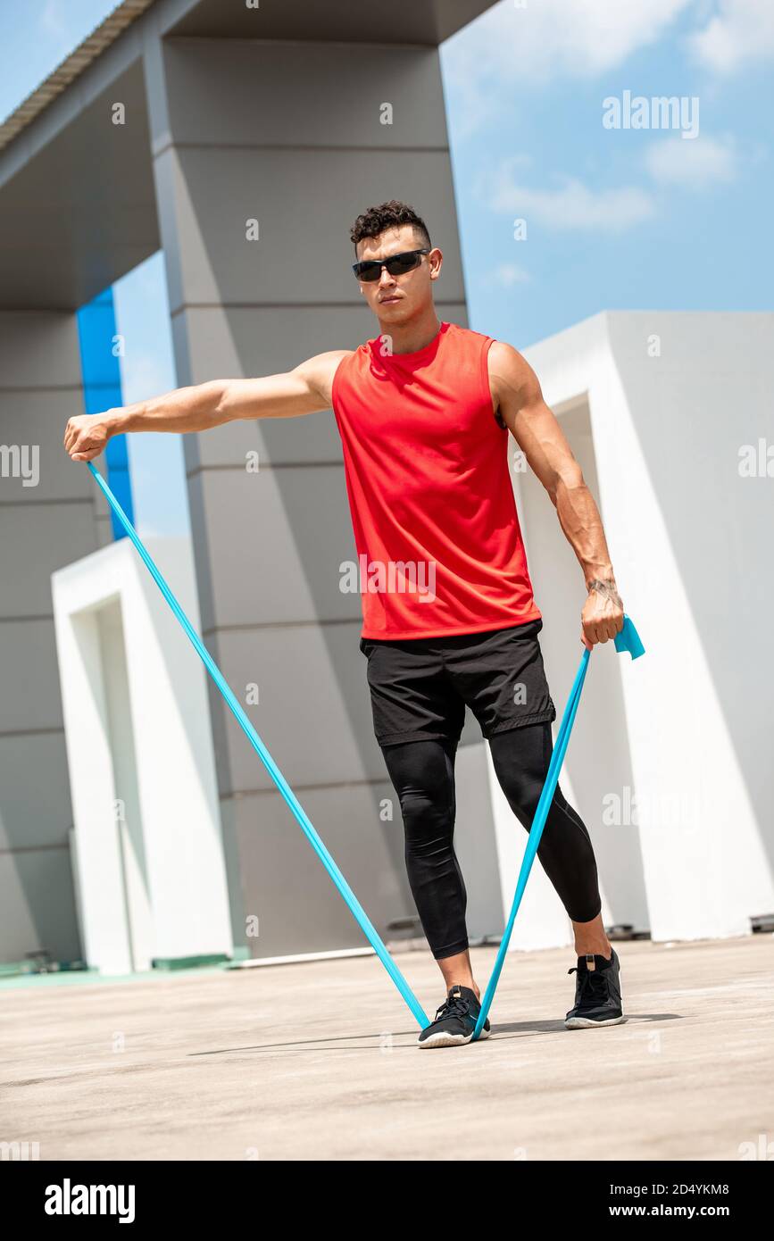 Handsome muscular sportsman doing shoulder lateral raise exercise with resistance band outdoors on rooftop Stock Photo