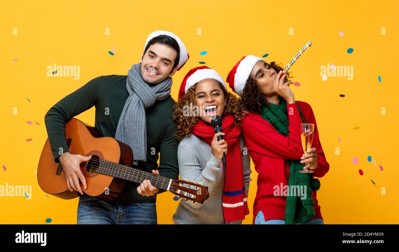 Happy three multiracial friends having party singing and celebrating Christmas in yellow studio background with confetti Stock Photo