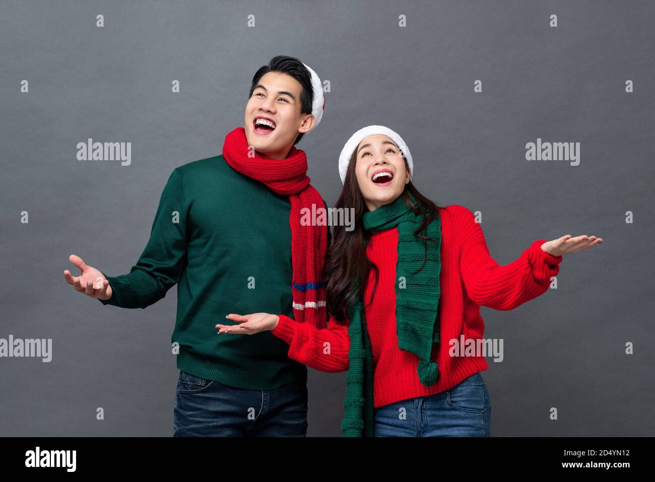Attractive Asian couple in Christmas attire smiling and looking up with hands open on isolated gray studio background Stock Photo
