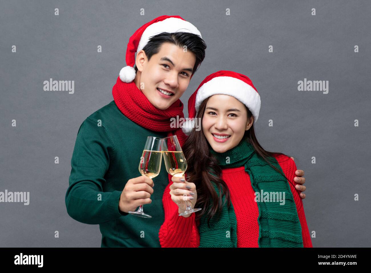 Smiling Asian couple in colorful red and green clothes drinking champagne celebrating Christmas in gray isolated background Stock Photo