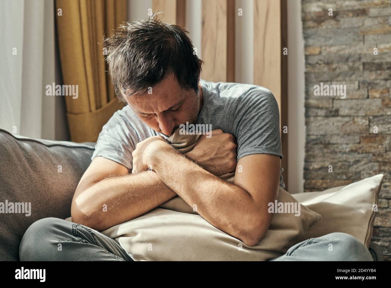 Depressed man sitting on living room sofa and hugging pillow, portrait of adult male having anxiety attack Stock Photo