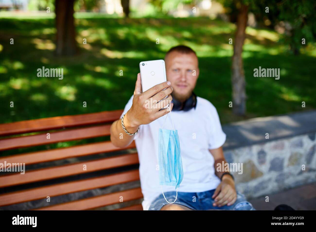 Man in medical mask sitting on the bench in the park take selfie by mobile phone. Male with respiratory protection is outdoors while talking and havin Stock Photo