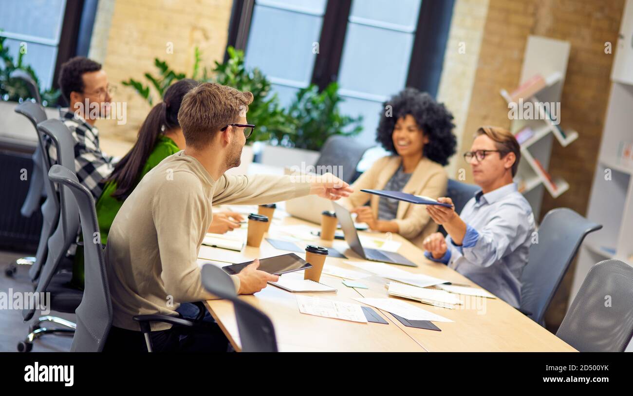 Caucasian man giving a folder with documents to his young colleague while having a meeting with project team in the modern office. Teamwork and collaboration, business people working together Stock Photo