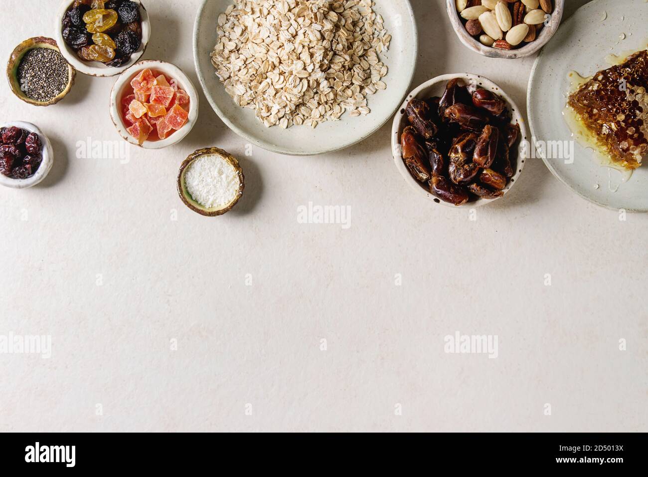 Variety of dried fruits, nuts, honey and oat flakes in ceramic bowls for cooking homemade healthy breakfast muesli or granola energy bars over white t Stock Photo