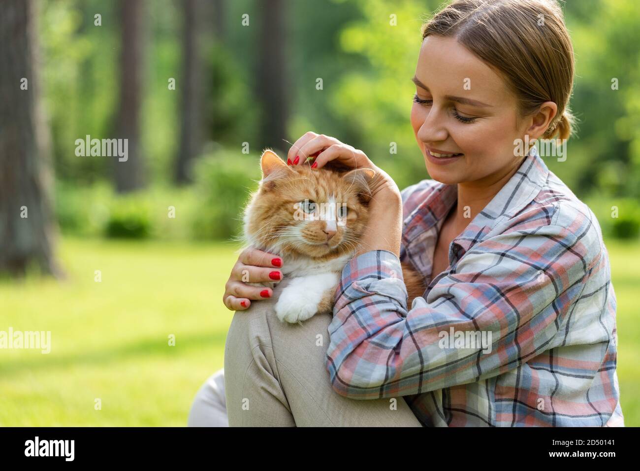 Close up of smiling woman in checked shirt hugging and embracing with tenderness and love domestic ginger cat, stroking on the head, outdoors in sunny Stock Photo