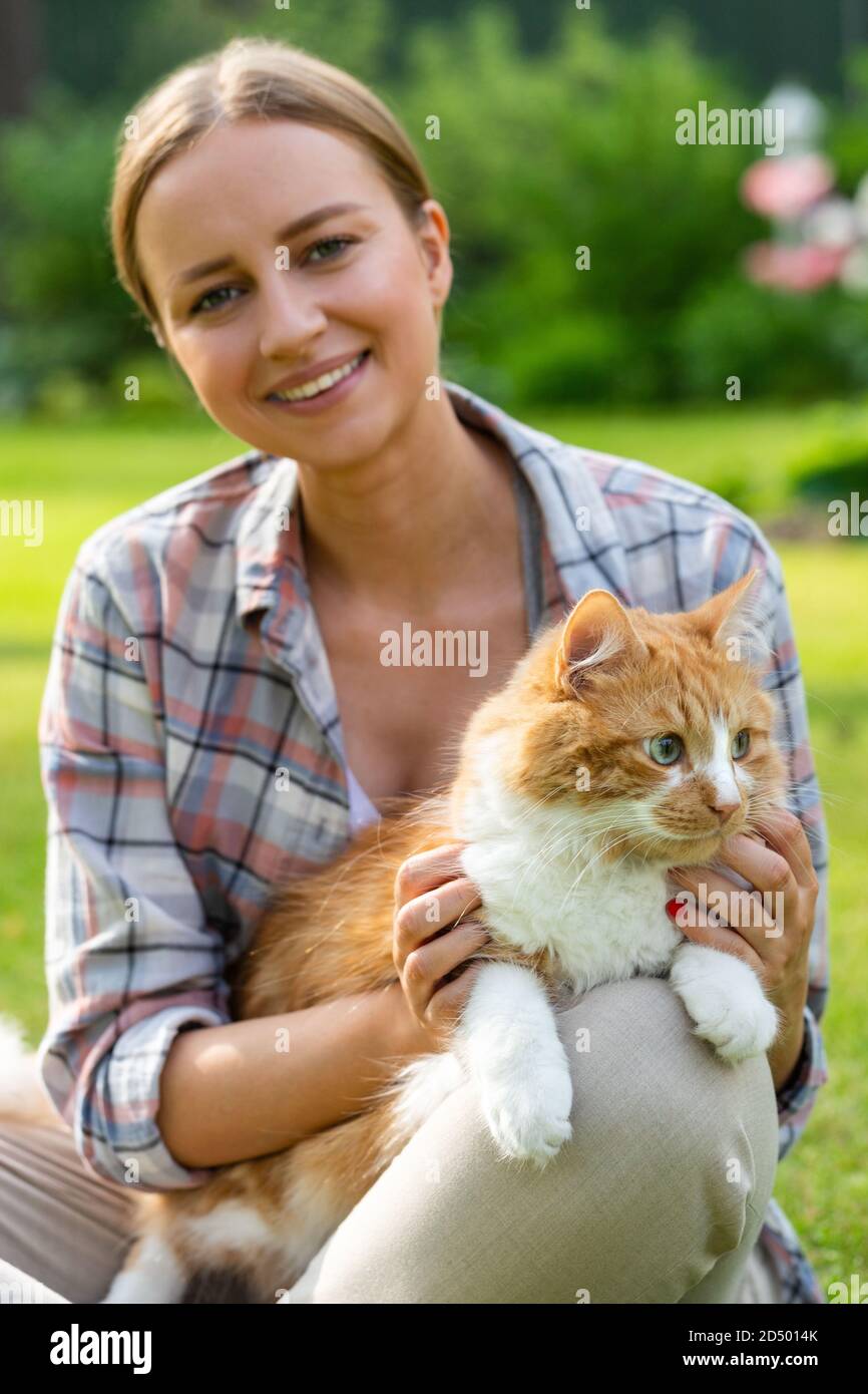 Close up of smiling woman in checked shirt looking at camera, hugging and embracing with tenderness and love domestic ginger cat outdoors. Love to the Stock Photo