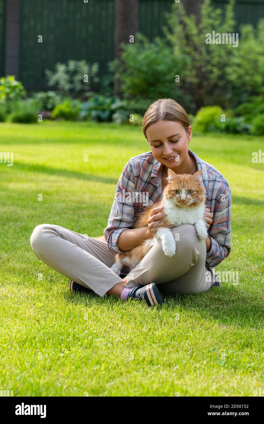 Close up of smiling woman in checked shirt hugging and embracing with tenderness and love domestic ginger cat, stroking on the head, outdoors in sunny Stock Photo