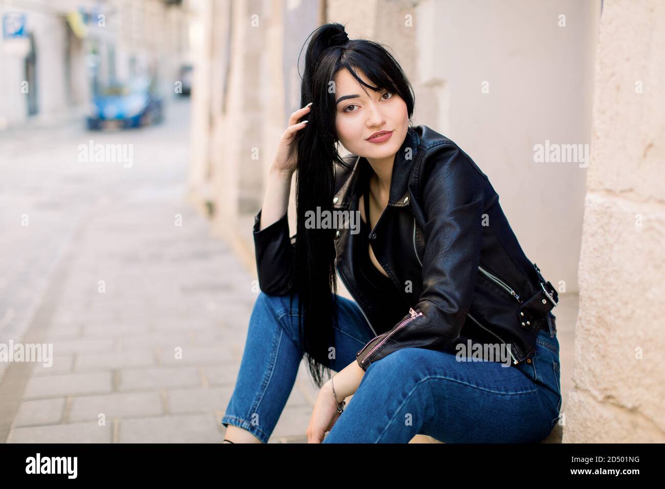 Portrait of young beautiful Asian lady in jeans pants and black leather jacket, sitting on the stone stairs near the old building in city. City Stock Photo