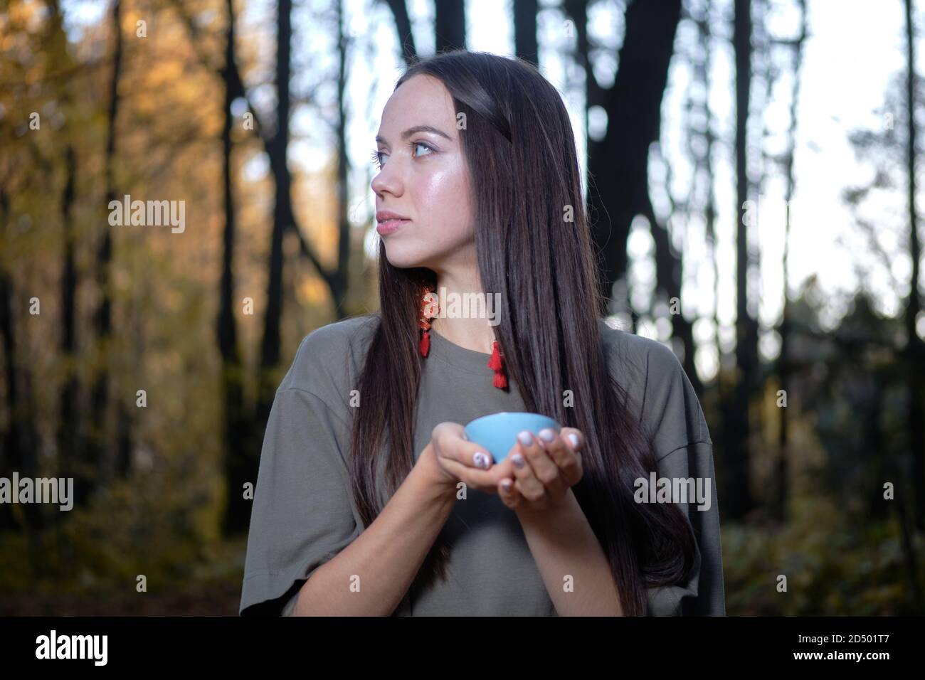 Young beautiful girl sitting on the ground in modern clothes in the autumn yellow forest and holding a tea ceremony Stock Photo