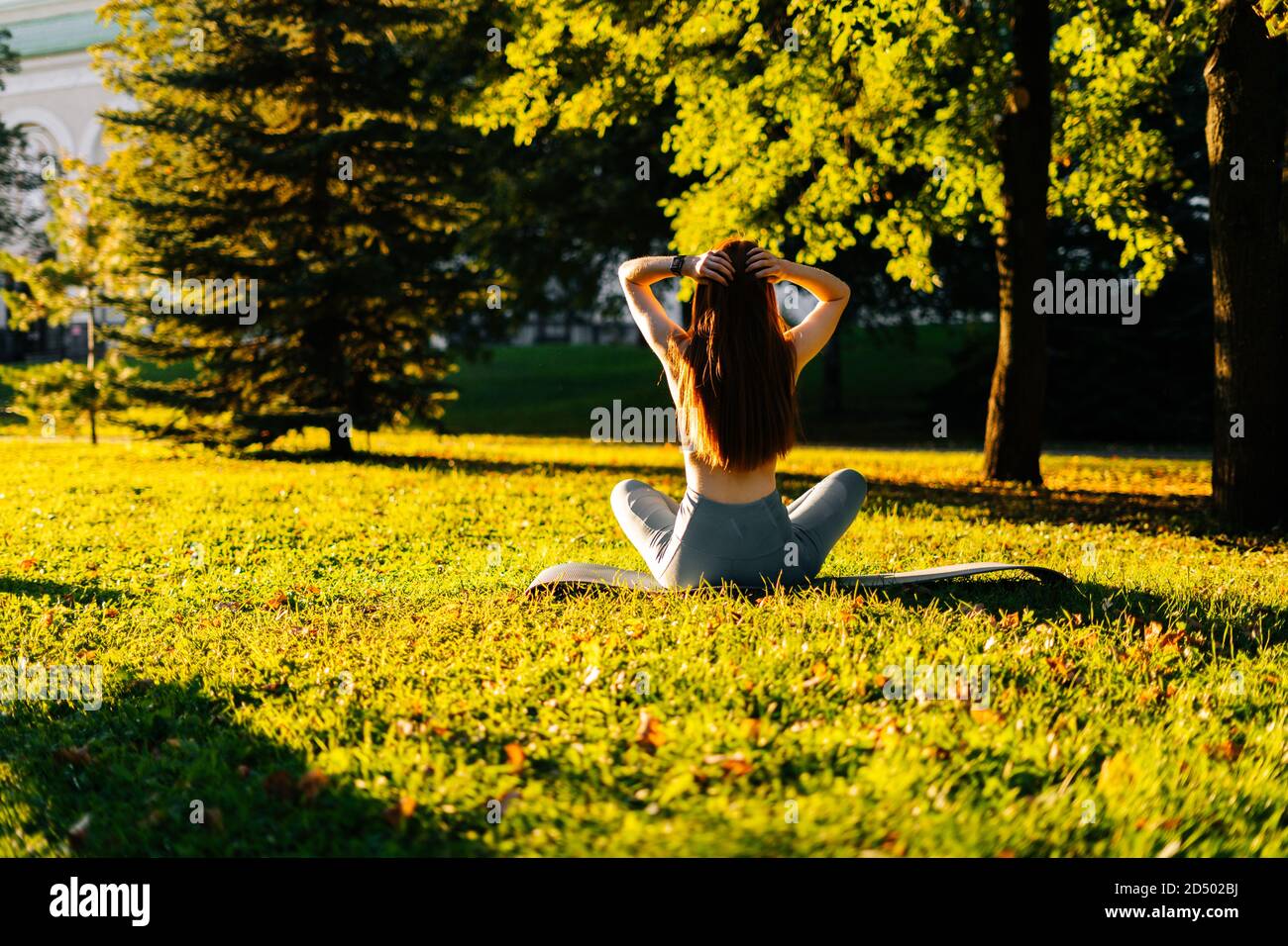 Back view of red-haired young woman sitting in lotus pose and hands up ...