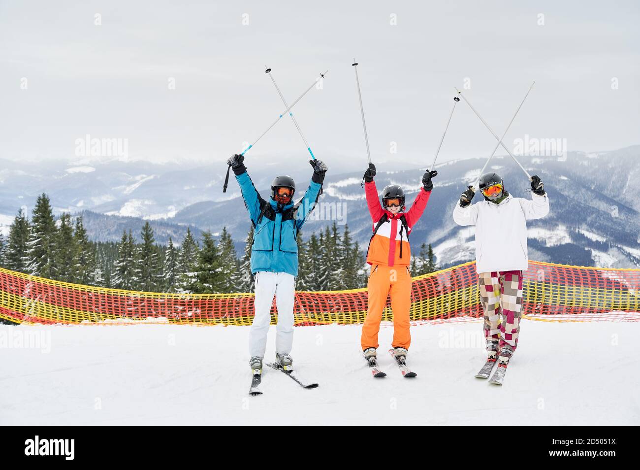 Three cheerful tourists posing on snowy slope against mountainous background, holding their ski poles up above. Friends at high spirits having fun at ski resort. Concept of winter sport and friendship Stock Photo