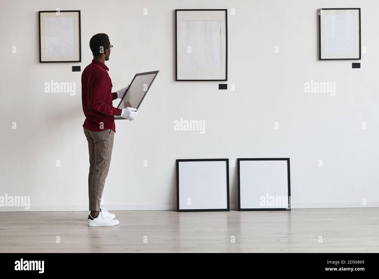 Full length portrait of African-American man planning art gallery or exhibition while setting up frames on white wall, copy space Stock Photo