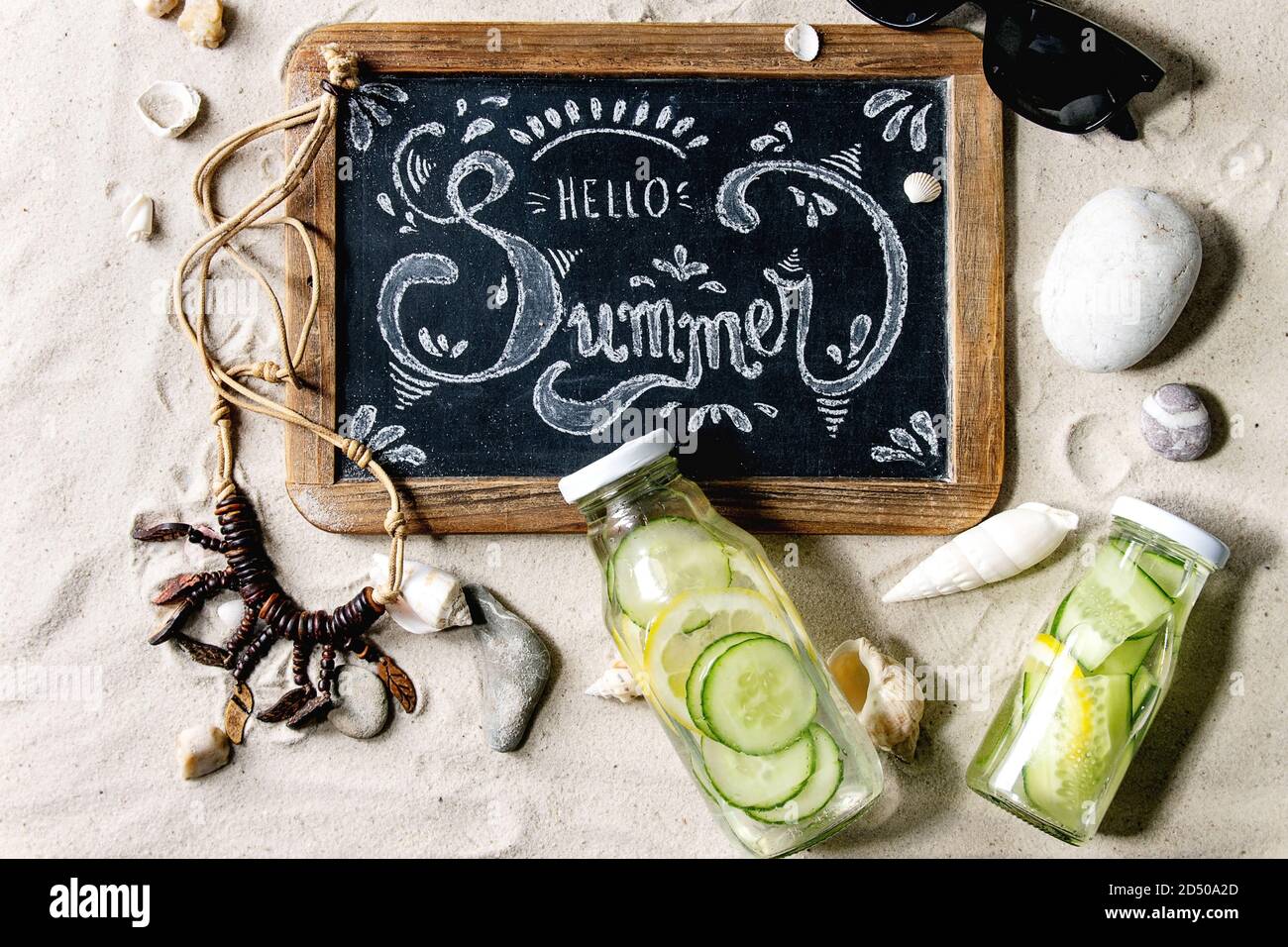 Summer theme. Chalk lettering Hello summer on vintage chalkboard, shells, sea stones, sunglasses, wooden beads, two glass bottles with sassy water on Stock Photo