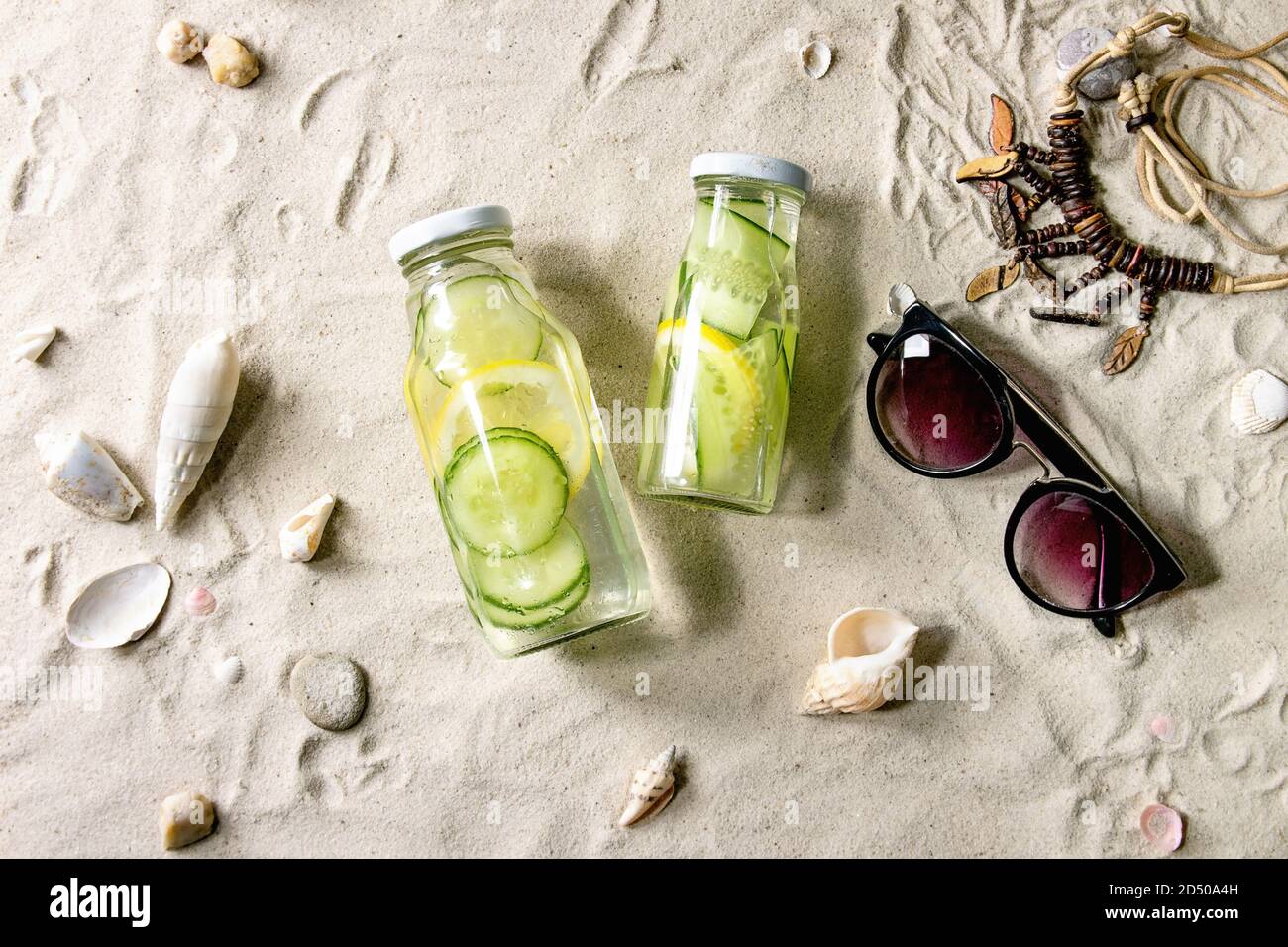 Summer theme. Two glass bottles with lemon and cucumber infusion sassy water, shells, sea stones, sunglasses, wooden beads, on white sand as backgroun Stock Photo