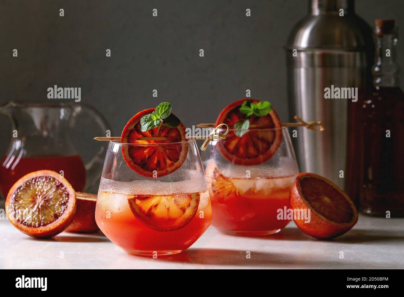 Blood orange iced cocktails in glasses, decorated by slice of oranges and fresh mint on skewers, served with shaker, jug of juice, bottle of rum on wh Stock Photo