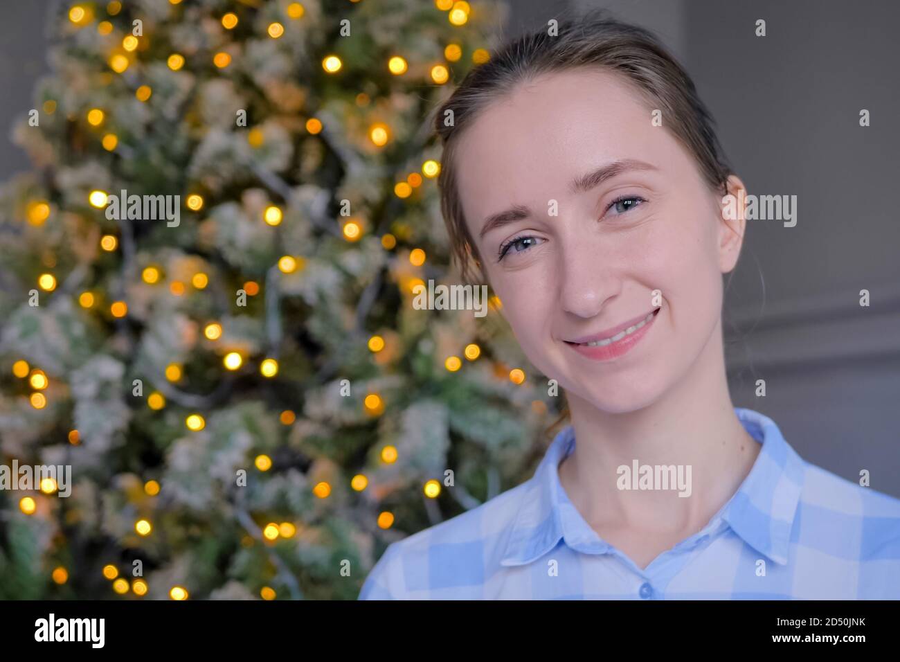 Portrait of young adult positive hipster, student or entrepreneur woman in plaid shirt smiling and looking at camera in room with Christmas tree Stock Photo