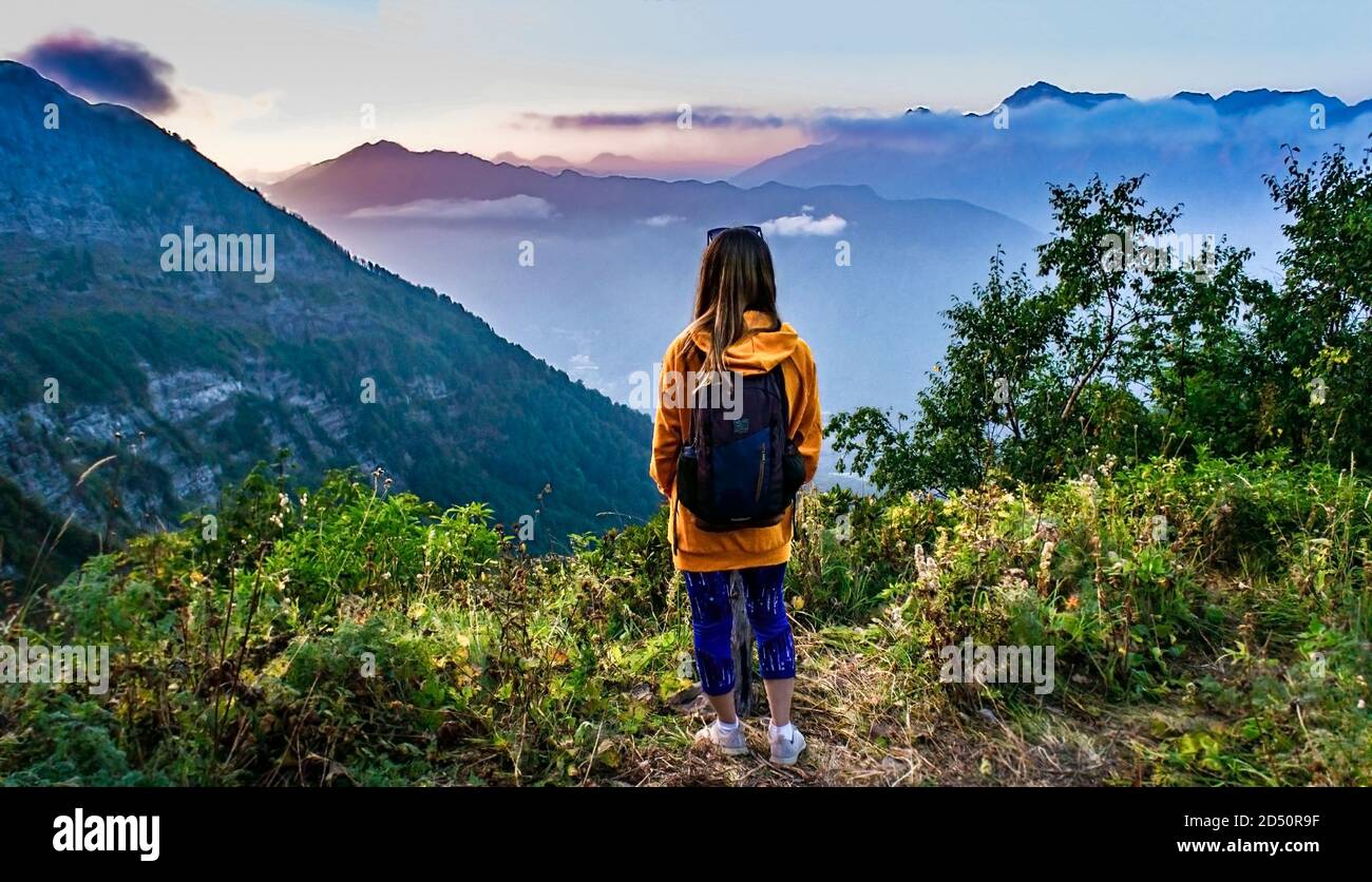 young blond woman from the back in yellow hoodie with a backpack looks at a bright sunset in the mountains. Trekking in mountains, active lifestyle, h Stock Photo