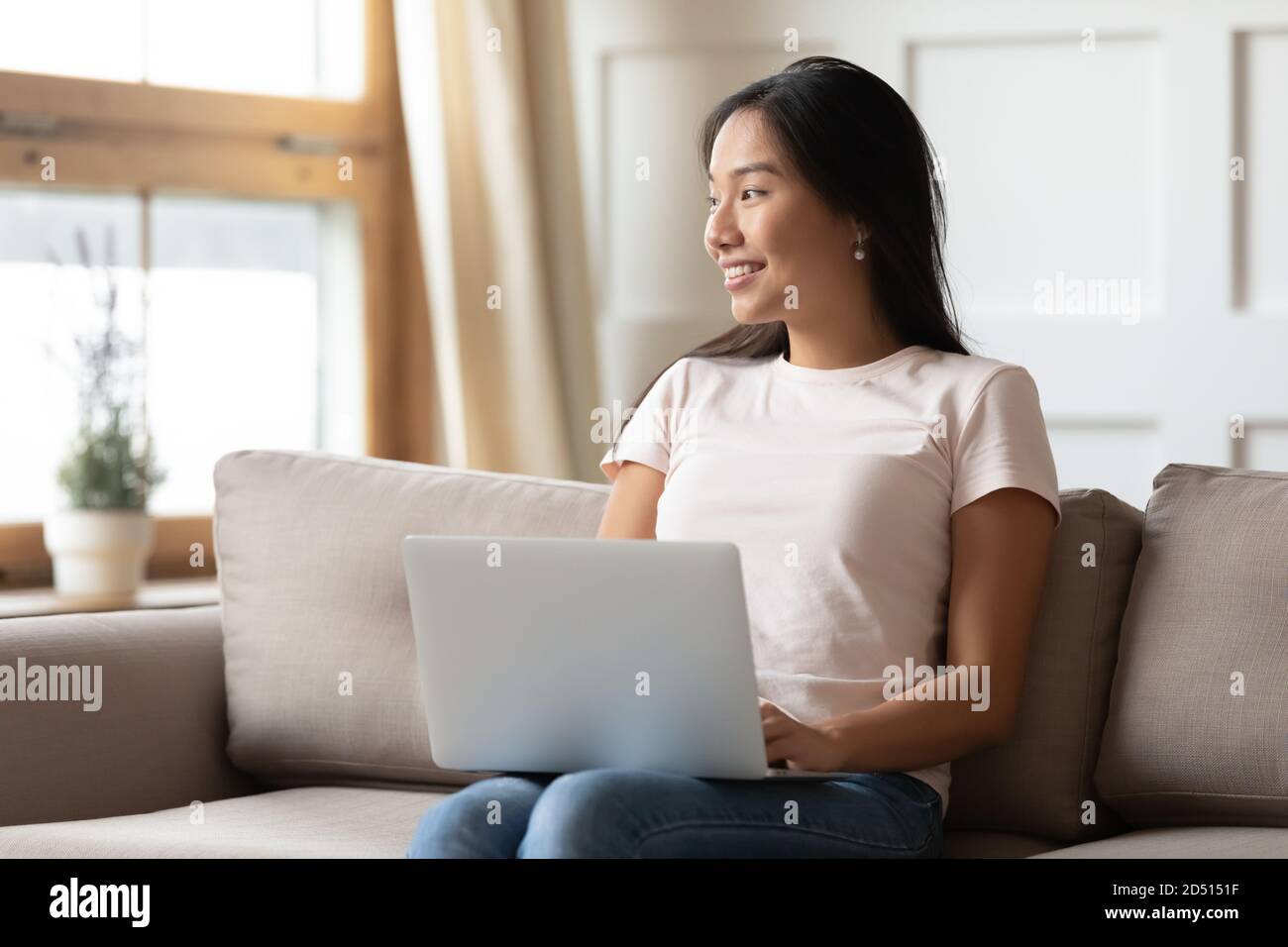 Smiling dreamy Asian young woman with laptop sitting on couch Stock ...