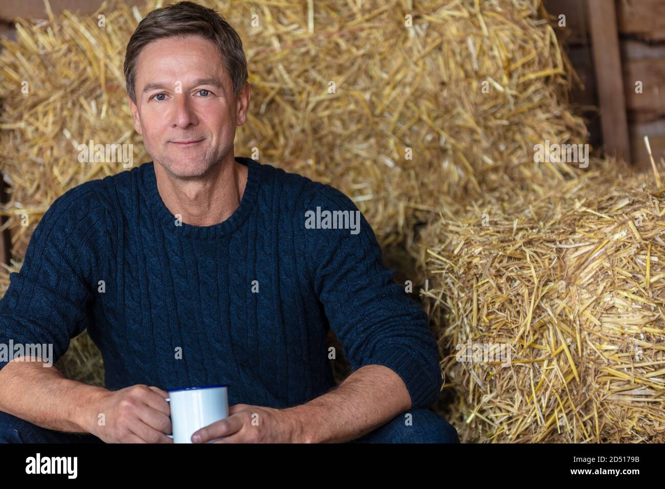 Portrait shot of an attractive, successful and happy middle aged man male wearing a blue sweater sitting on hay bales in a barn or stables drinking cu Stock Photo