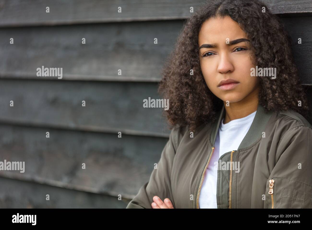 Beautiful mixed race African American girl teenager female young woman outside wearing a green bomber jacket looking sad depressed or thoughtful Stock Photo