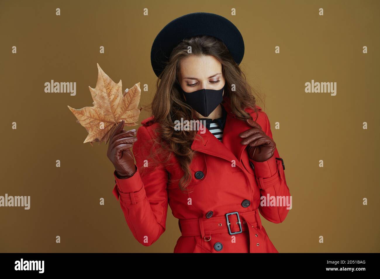 Life during covid-19 pandemic. elegant female in red coat with black mask and yellow autumn maple leaf isolated on beige background. Stock Photo