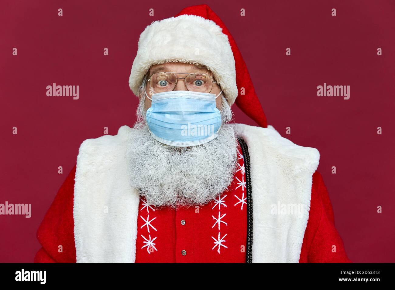 Funny bearded Santa Claus wearing face mask looking at camera on red background. Stock Photo