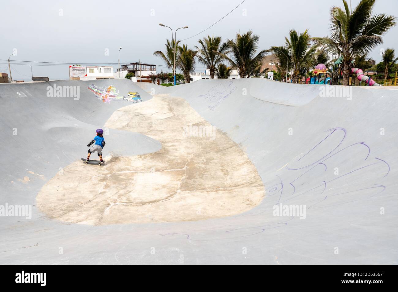 A young boy wearing trendy clothes, a purple helmet, and a face mask, skates in SKATEPARK SAN BARTOLO POZAS BOWLS during the pandemic period. Stock Photo