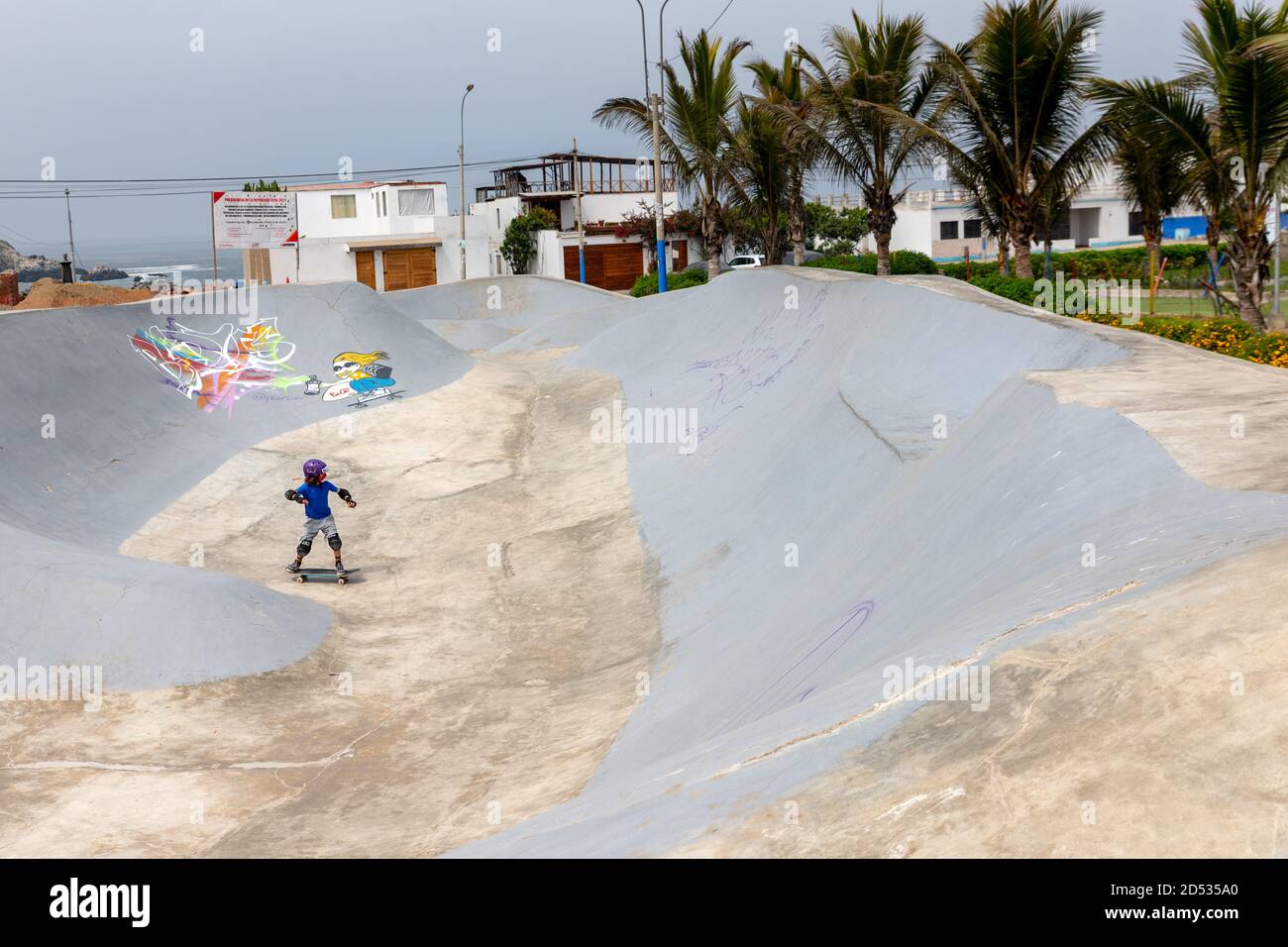 A young boy wearing trendy clothes, a purple helmet, and a face mask, skates in SKATEPARK SAN BARTOLO POZAS BOWLS during the pandemic period. Stock Photo