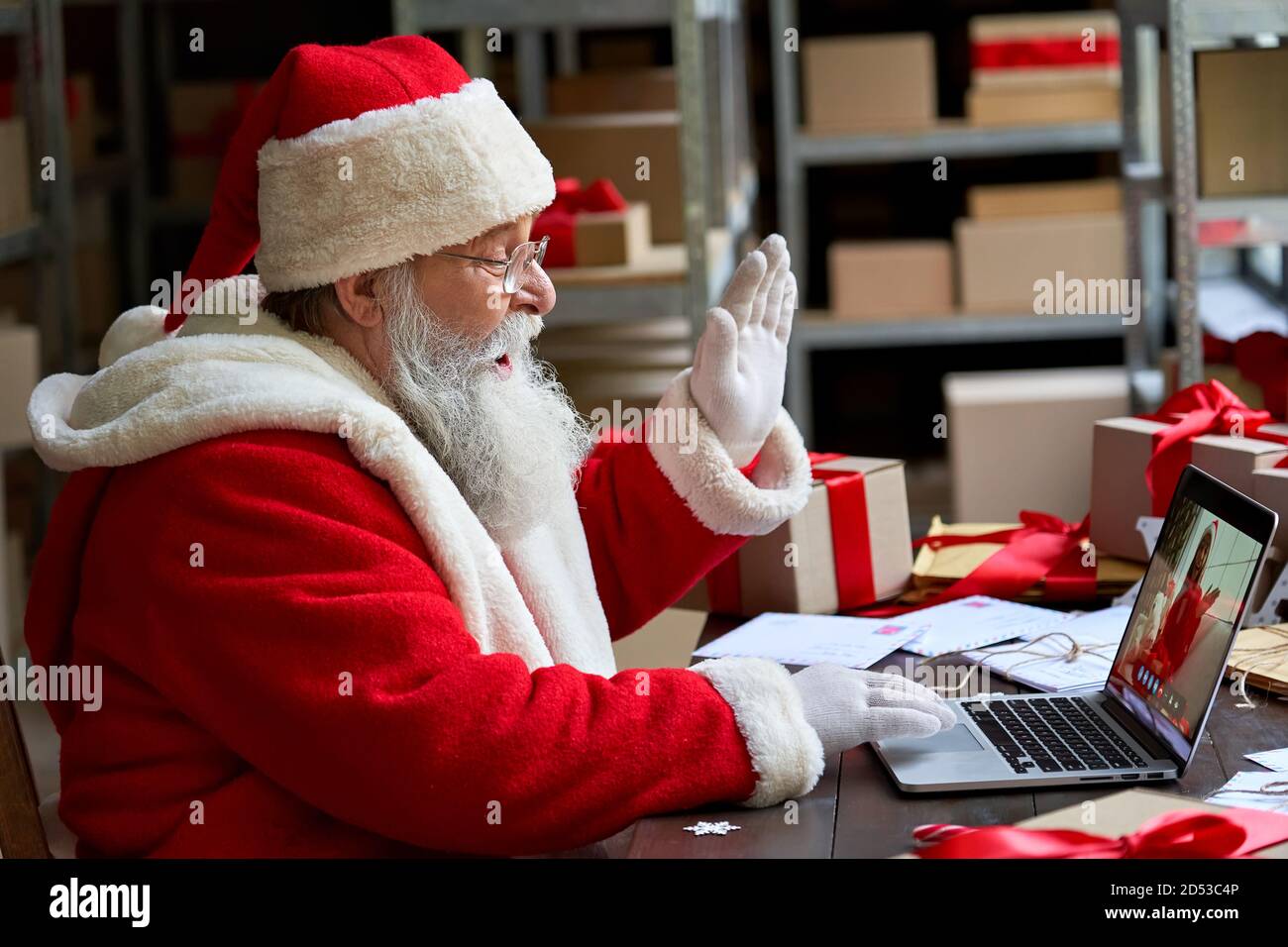 Santa Claus video calling on laptop greeting child sit at workshop table. Stock Photo