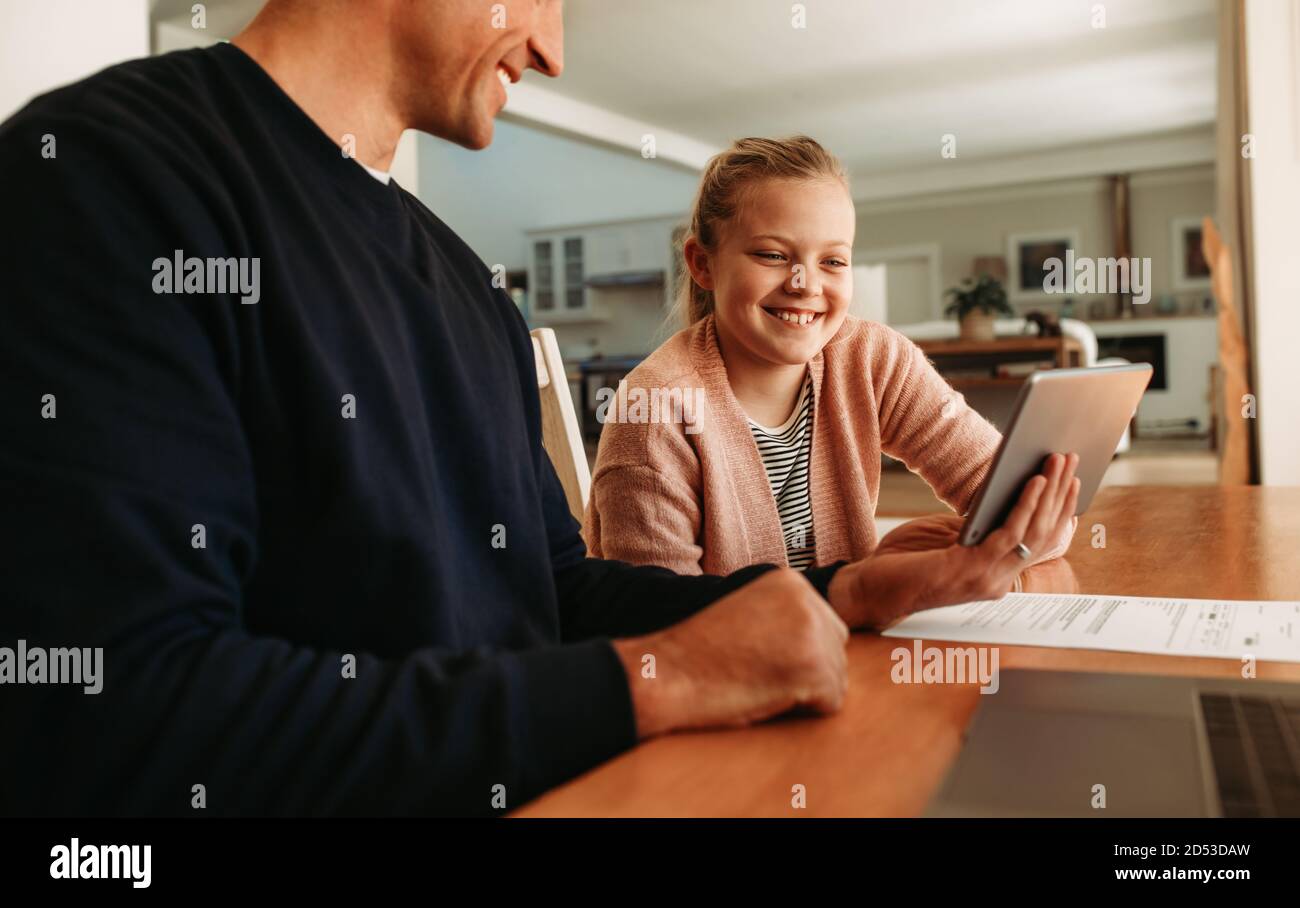 Father and daughter sitting at table using a digital tablet together at home. Girl looking at tablet computer in hand of her father at home. Stock Photo