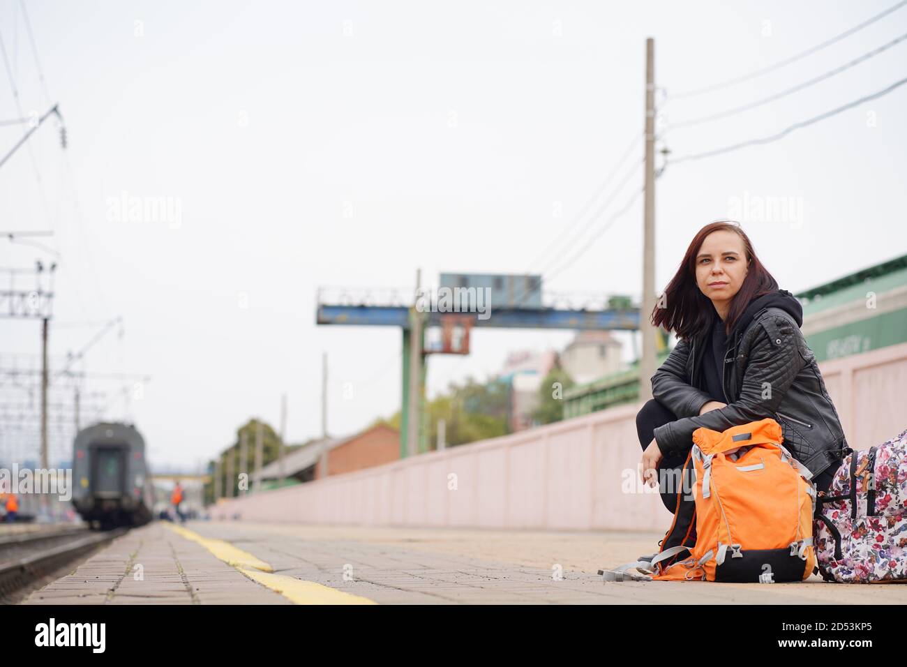 Young woman squats on platform, waiting for train. Female passenger with backpacks sitting on railroad platform in waiting for train ride. Stock Photo