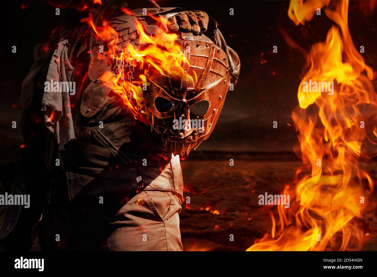 Photo of a post apocalyptic raider warrior in leather jacket with metal armor  standing in night wasteland and holding a burning metal mask. Stock Photo