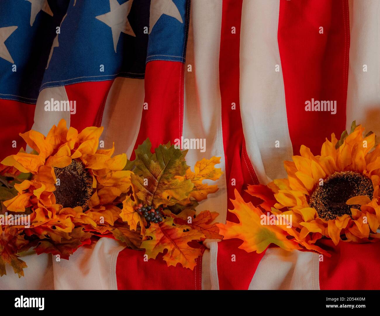 Sunflowers and fall leaves sit on on American flag that is draped and glows in the warm light. Stock Photo