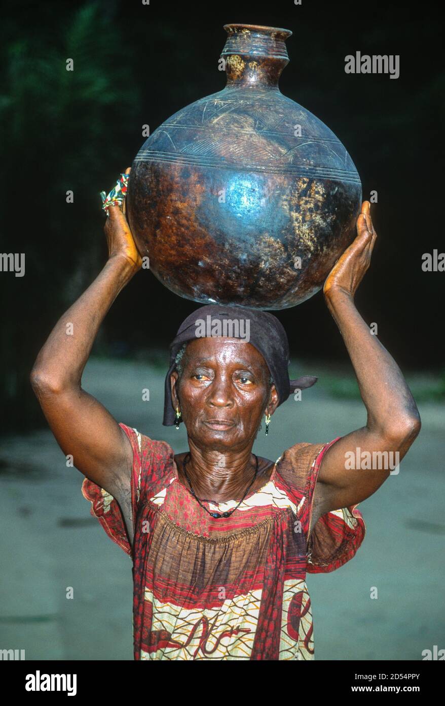 Ouassou, Ivory Coast. Elderly Ivorian Woman Carrying Pot on Head. Stock Photo