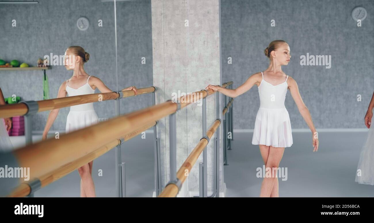 Cute girl dancer in white costume does exercises leaning on wooden barre near large mirror in light studio at ballet training Stock Photo