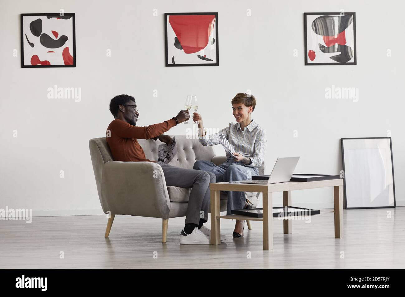 Portrait of young mixed-race couple enjoying champagne while sitting on couch in room decorated by modern art, copy space Stock Photo