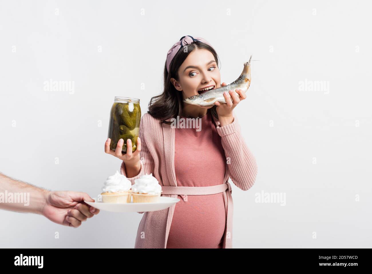 pregnant woman holding jar with pickled cucumbers and eating dried fish while looking at cupcakes on plate in male hand isolated on white Stock Photo