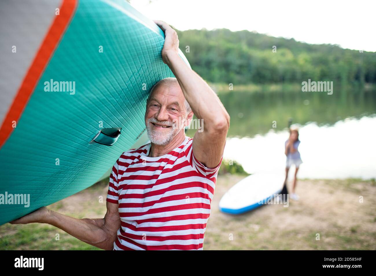Senior man carrying paddleboard by lake in summer, looking at camera. Stock Photo