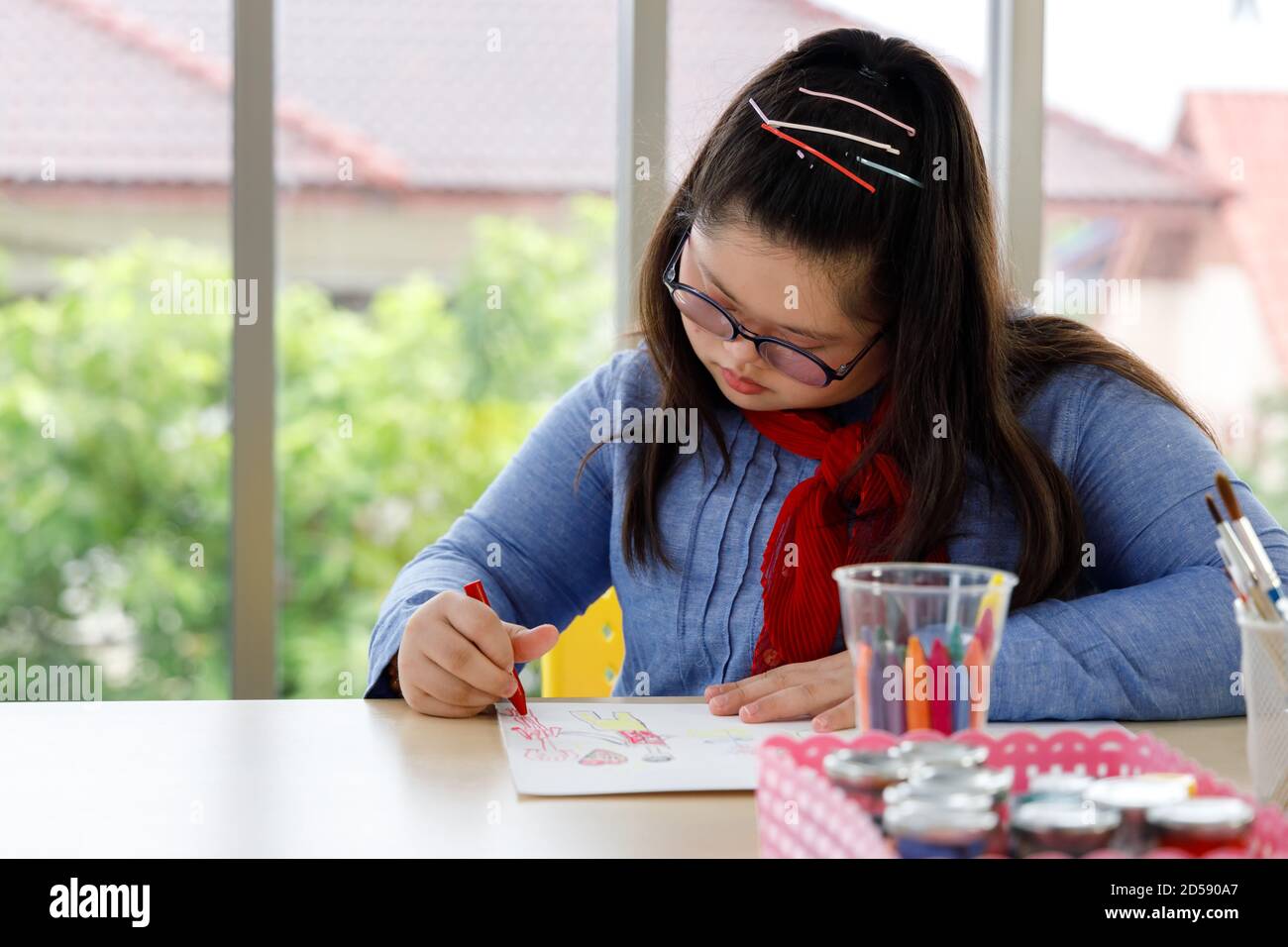 Girl with Down's syndrome drawing picture with crayon in art class. Concept disabled kid learning. Stock Photo