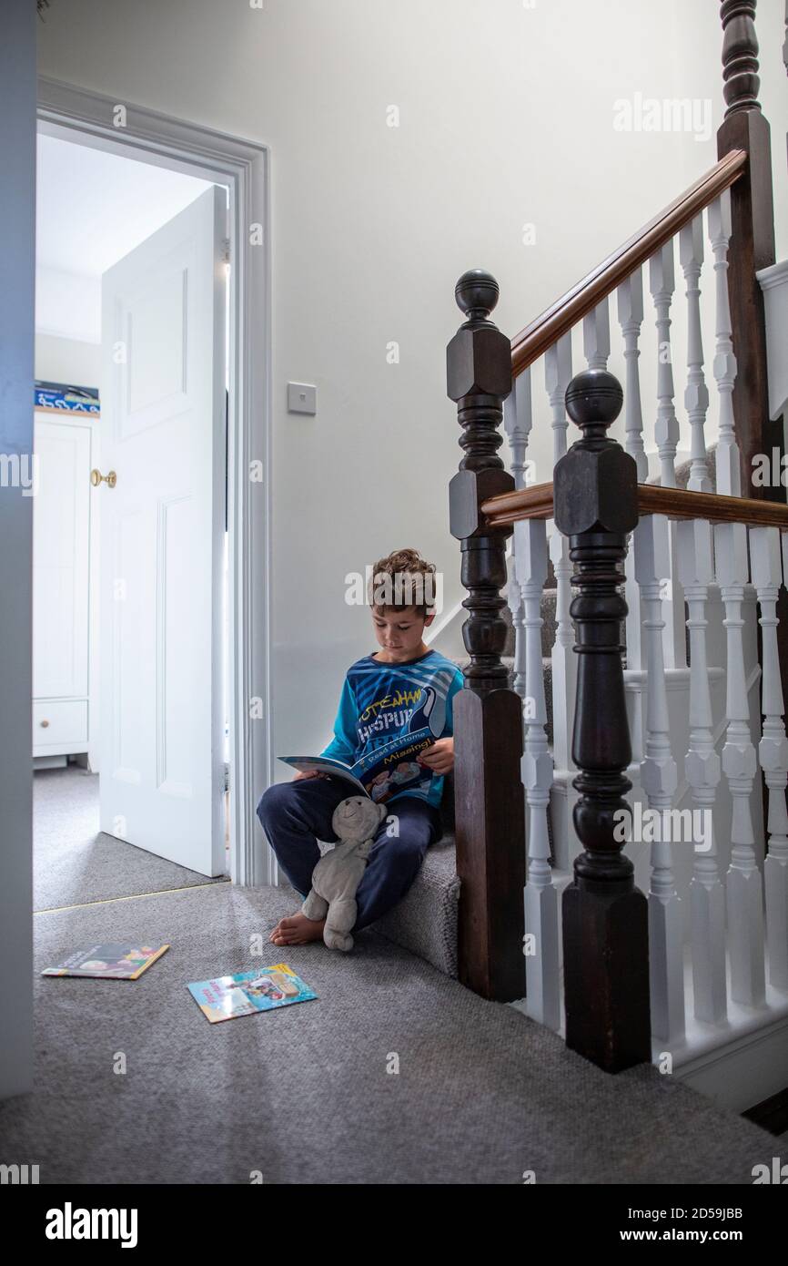 Six year old boy reading a book whilst sat on his residential home staircase, England, United Kingdom Stock Photo