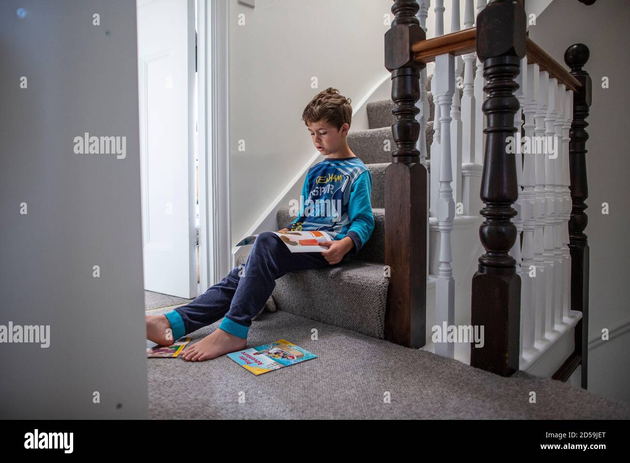 Six year old boy reading a book whilst sat on his residential home staircase, England, United Kingdom Stock Photo